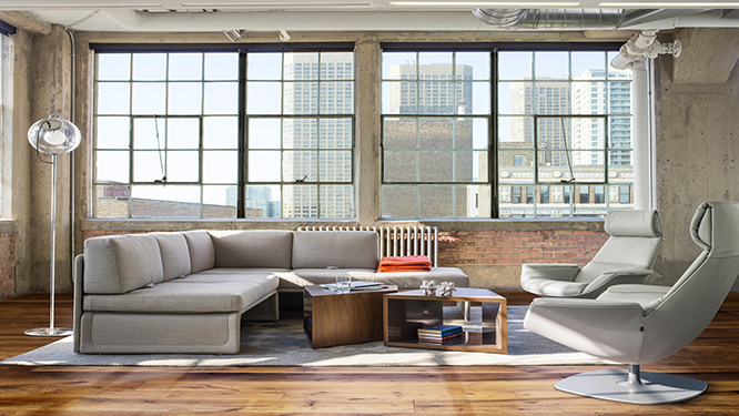 Large grey sectional, two brown cubed coffee tables, two grey lounge chairs and a grey rug in collaborative space with windows looking out to the city.