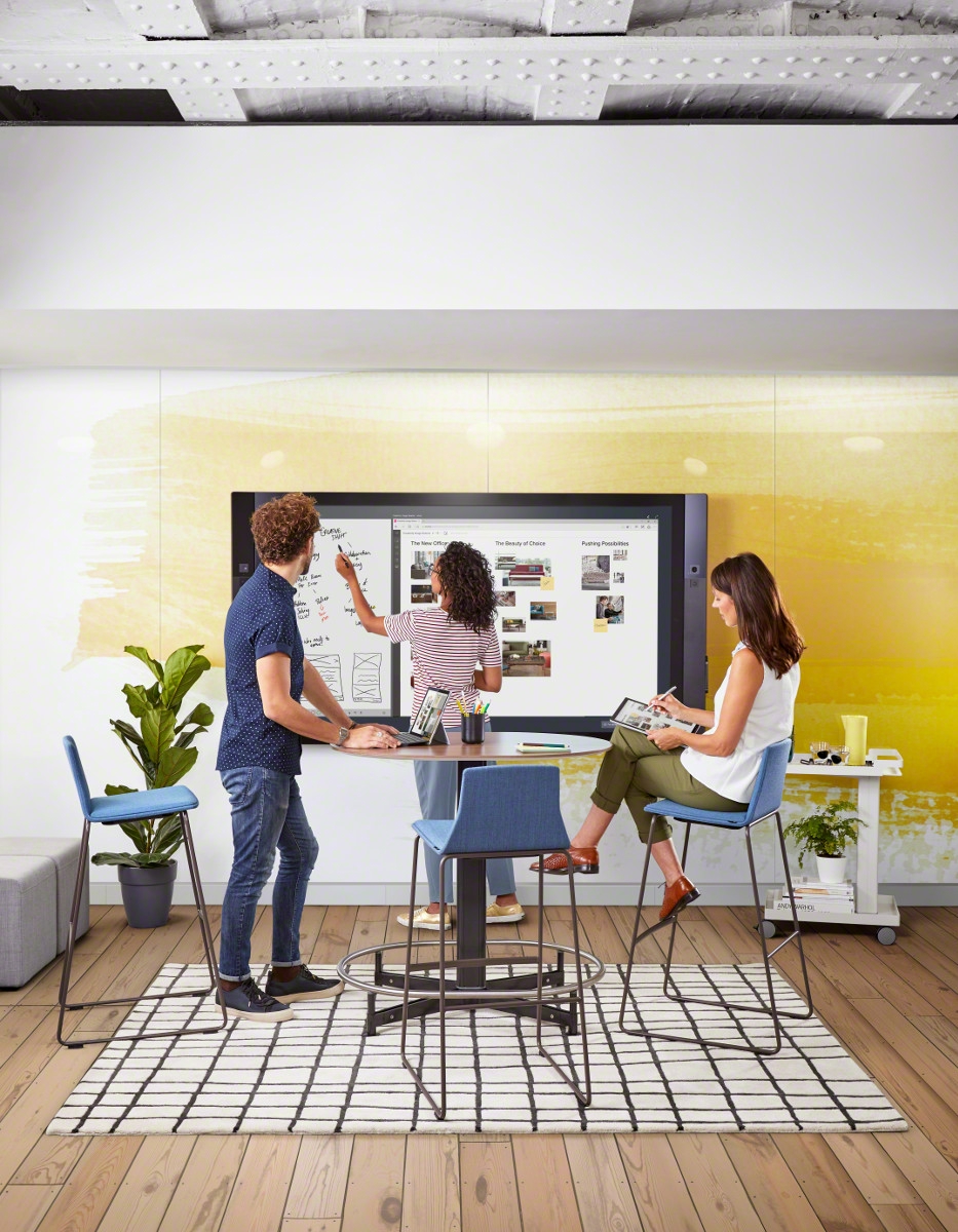 Woman sits on stool at standing-height table, as another man and woman stand and collaborate on Microsoft Surface Hub technology