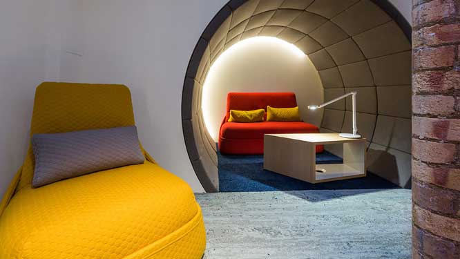 Yellow lounge chair with grey pillow and red lounge sofa with wooden coffee table and table light in small circular relaxation room.