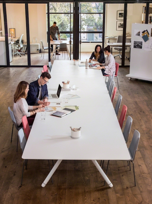 People collaborating at long, white conference table with glass door and windows leading to private meeting room in the background.