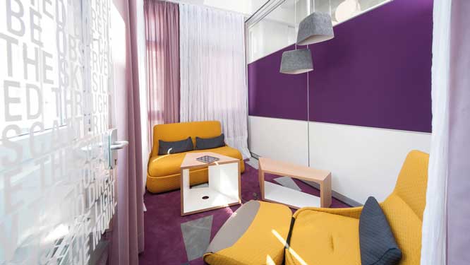Small relaxation room with yellow sofa and lounge chair, two wooden side tables, two hanging grey lights and purple wall, carpeting and curtains.