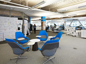 Informal office gathering space with blue chairs and small round table near meeting room and work areas