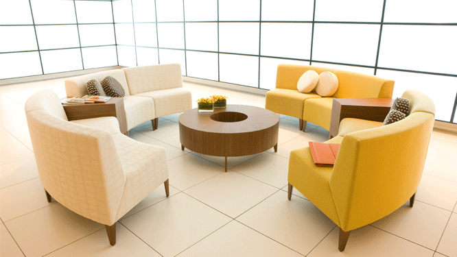 A collaborative room with cream colored sofas, yellow sofas and a round, wooden coffee table on tiled floors with lighted, tiled walls.