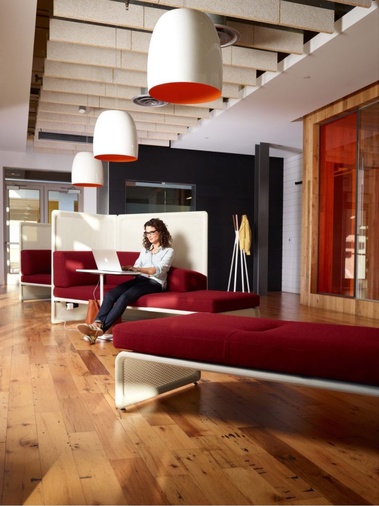 Woman sitting at laptop in office lounge space with red upholstered bench seating, white privacy walls, and matching ceiling lamps