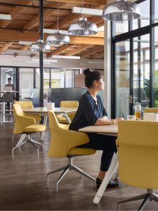 Woman looking out of a window in office cafe space, seated on yellow lounge chair at wooden table