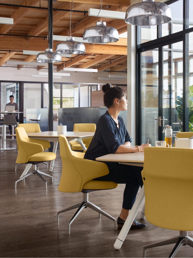 Woman looking out of a window in office cafe space, seated on yellow lounge chair at wooden table