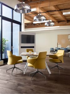 Square wooden office meeting table with yellow chairs, mobile wall with monitor, and hanging metal lamps