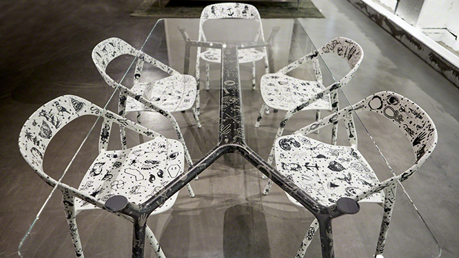 Long glass top table with metal black legs and five white dining chairs with tattoo art covering them.