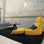 Living room with a concrete wall, featuring a yellow lounge chair with an attached footrest on a black rug, a blue cushion and electronic tablet on the chair, a round wooden coffee table with decorative items, a beige ottoman with stacked books, a tall floor lamp, and a large abstract painting on the wall.