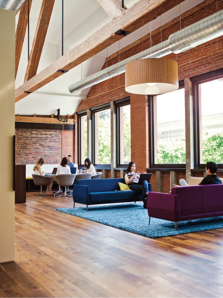 Social space in office with colorful plush couches and chairs, matching tables, hanging lamp, and bench seating against wall