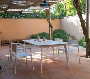 Outdoor patio with white outdoor chairs around a rectangular table and greenery surrounding the sitting area