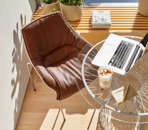 Overhead view of brown leather lounge chair, vase shaped and glass topped table with laptop and small stool with blue cushion on a balcony