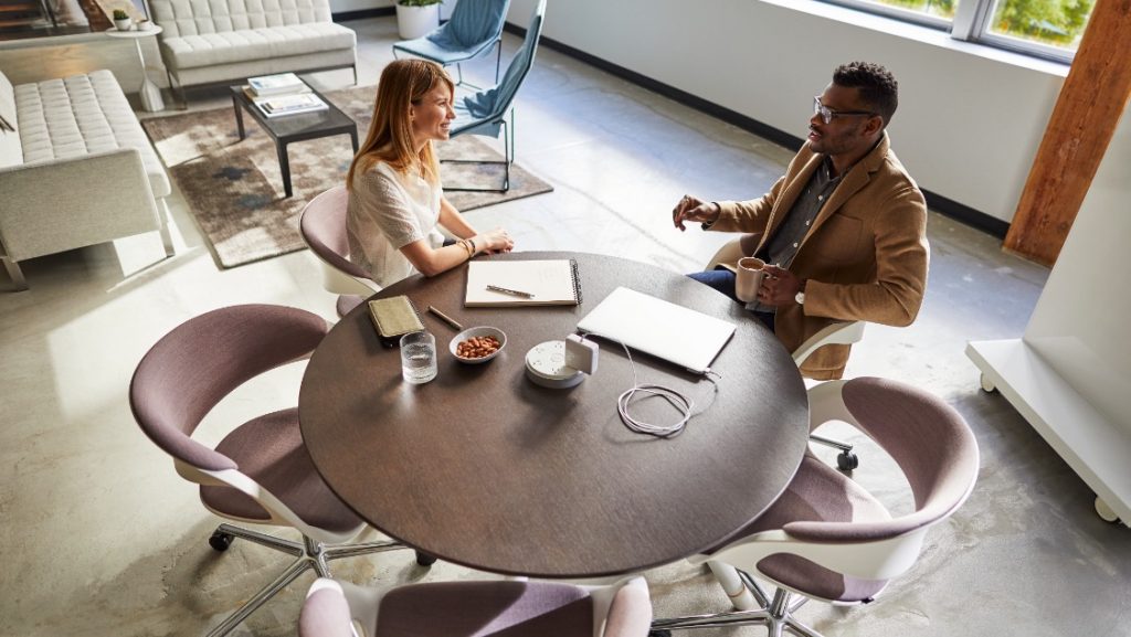 Two workers in office collaborating around round coffee table with notebooks and laptop