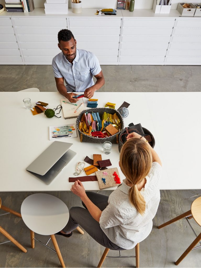 Two workers handling fabric samples at a white meeting table with laptop, drawing supplies, and cabinet behind them
