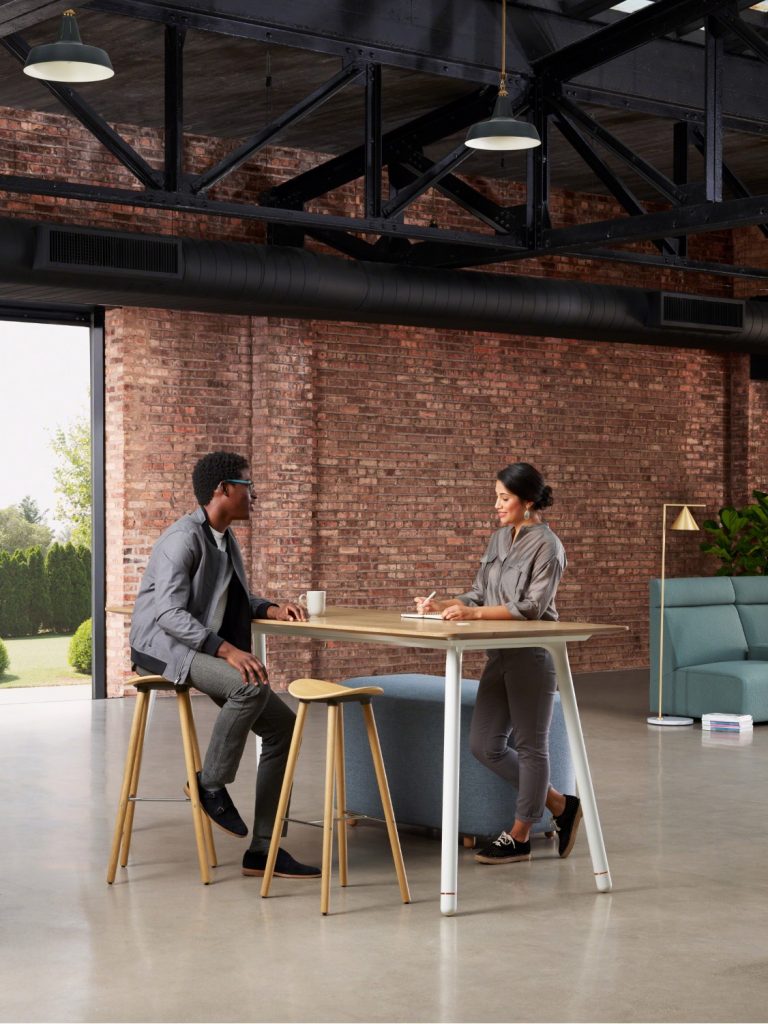 Two young office workers sitting at collaborative table in modern office building with table, bench, and stools