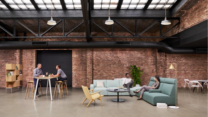 Office area with industrial ceiling and brick walls along with employees gathered at pastel colored couches and standing-height table with stools.