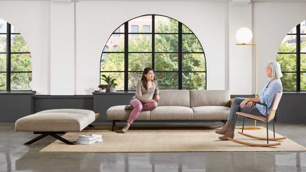 Coworkers sitting on a rocking chair and grey office sectional couch in front of arched window