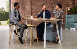 Three young office workers around wooden top meeting table inside office common area