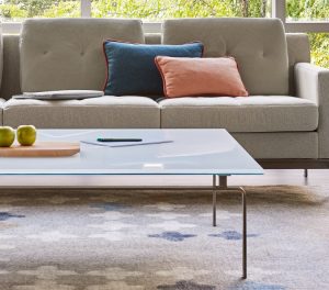 Glass top coffee table on grey patterned rug with grey office couch nearby