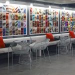 Seating area featuring a long gray bench with red pillows, round glass tables, and wireframe chairs, set against a vibrant wall art installation made of colorful abstract images arranged in a grid.