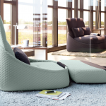 Living space with a light green lounge chair with an adjustable backrest and a small black pillow. A wooden side table with a white lamp and a cup is beside it, and a dark gray lounge chair is visible in the background.