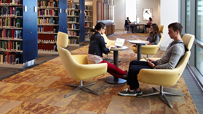 College library with students sitting in yellow lounge chairs with small individual laptop tables.