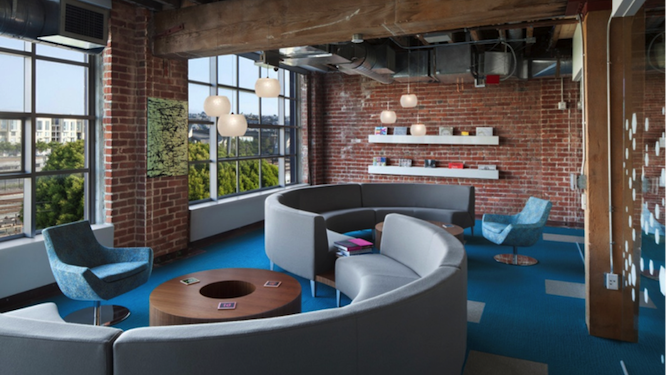 Large meeting room with red brick walls, two grey lounge sofas and blue lounge chairs around two big round wooden coffee tables