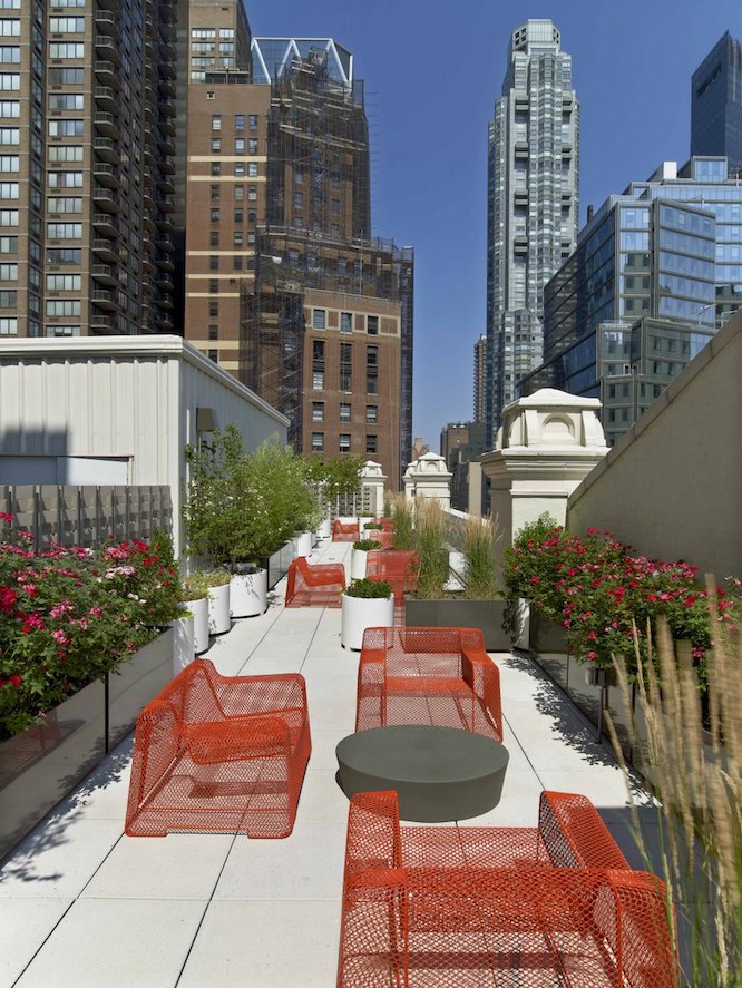 Outdoor, rooftop sitting area with red wired chairs, low black coffee tables and decorative greenery with tall city buildings in the background.