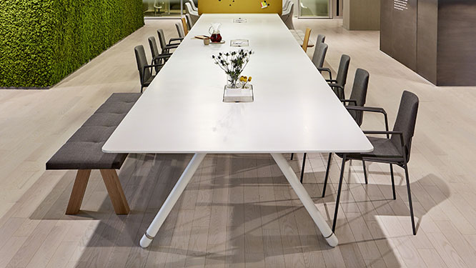 Large white conference table with black office chairs and black cushioned bench on light hardwood floors.
