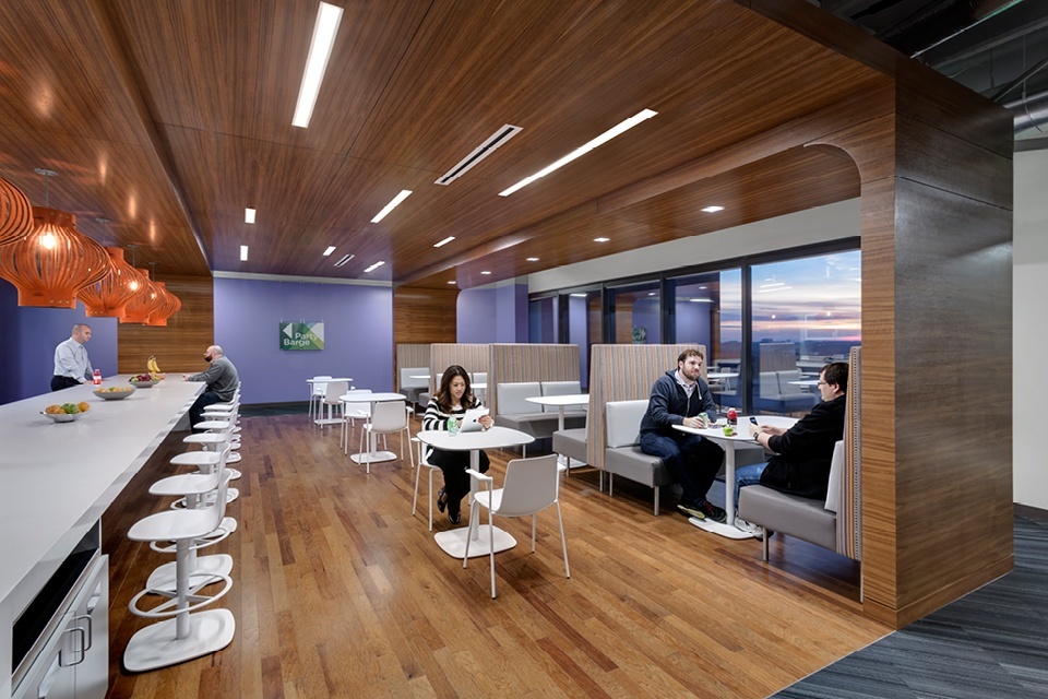 Open work space with bar style counters with white bar stools, small white tables with white office chairs and grey booths with white tables on a hardwood floor.