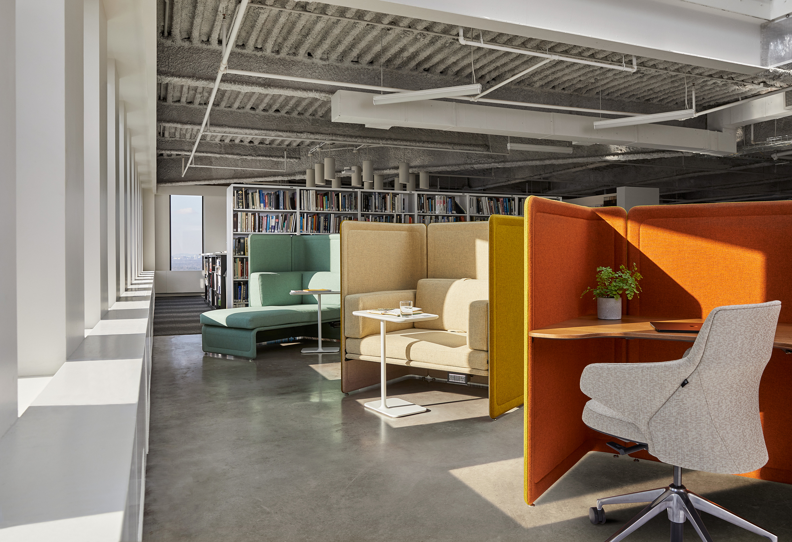 Modern workspace with high‑back lounge seating in green, beige, and orange tones, arranged among bookshelves and small tables