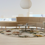 open space with wooden flor, circular rug and large windows with curtains. In the center we see a light brown circa lounge system, a coffee table and personal table on the side