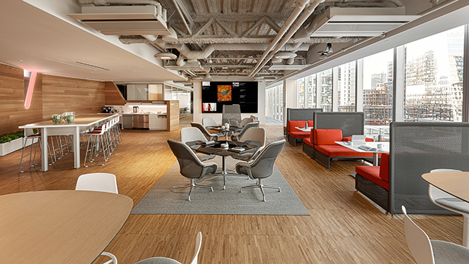 Cafe style work space with wooden floors, a large conference table with bar stools, small work tables with grey lounge chairs and restaurant style orange booths with large white tables.