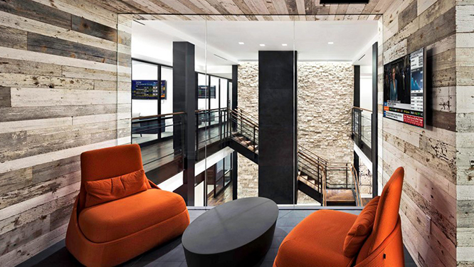 Dark orange lounge seating with oval shaped coffee table with wall mounted TV on wood paneled walls and large window overlooking office.