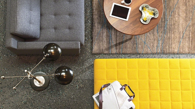 Look down view of a grey couch, bright yellow ottoman, small round wooden coffee table and black standing lamp with brown rug.