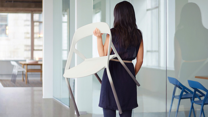 Woman in dark blue dress walking down hallway holding a lightweight white office chair.