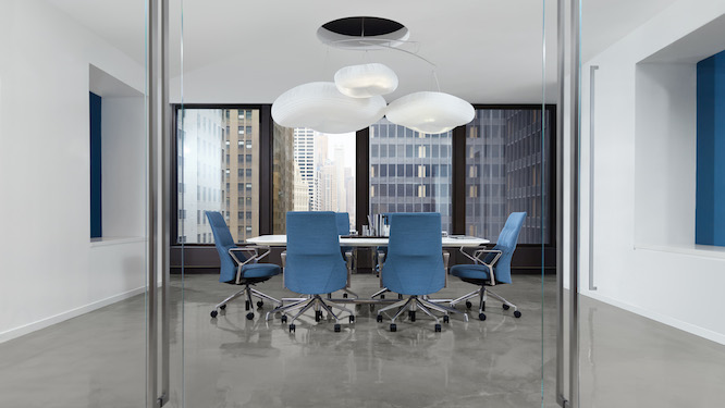 Large white conference table with blue lounge conference chairs and large hanging white lights in a large conference room with large floor to ceiling windows.