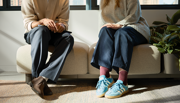 Two people seated on a cushioned bench, with only their lower legs and shoes visible in natural light.