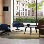 Modern lounge area with blue chairs, upholstered seating, round table, and floor lamp near large windows