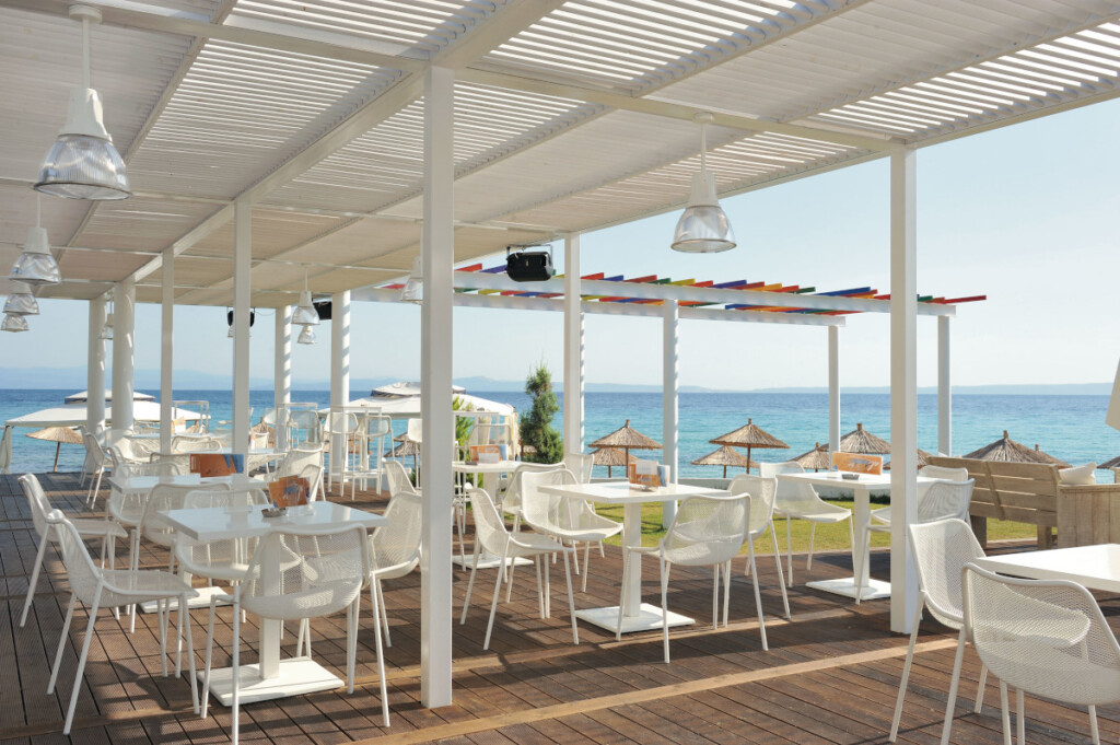 Outdoor café seating with white tables and chairs under a pergola, overlooking the sea
