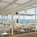 Outdoor café seating with white tables and chairs under a pergola, overlooking the sea