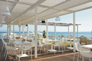 Outdoor café seating with white tables and chairs under a pergola, overlooking the sea