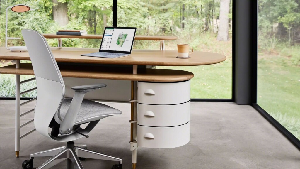 Modern home office with a curved wooden desk, chair, laptop, and storage drawers, set beside large windows overlooking greenery.