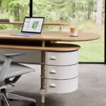 Modern home office with a curved wooden desk, chair, laptop, and storage drawers, set beside large windows overlooking greenery.