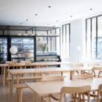 Minimalist café interior featuring light wood tables and chairs with refrigerated displays in the background.