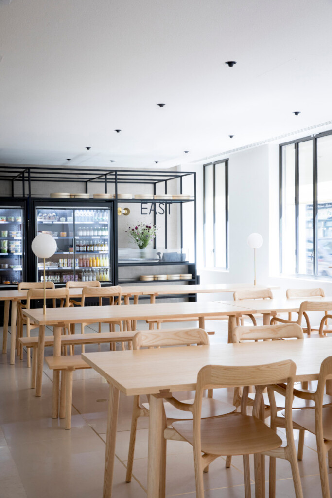 Minimalist café interior featuring light wood tables and chairs with refrigerated displays in the background.