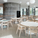 Bright café interior with round tables, light wood chairs, concrete columns, and hanging globe lights.