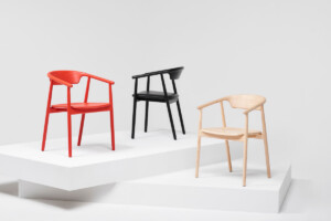 Three modern armchairs displayed on white platforms in red, black, and natural wood finishes