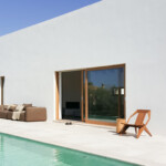 Modern white house exterior with a pool, patio seating, and wooden lounge chair under a clear blue sky