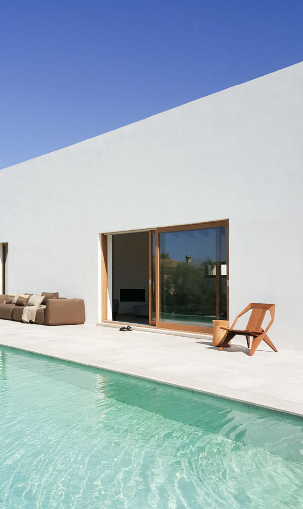Modern white house exterior with a pool, patio seating, and wooden lounge chair under a clear blue sky